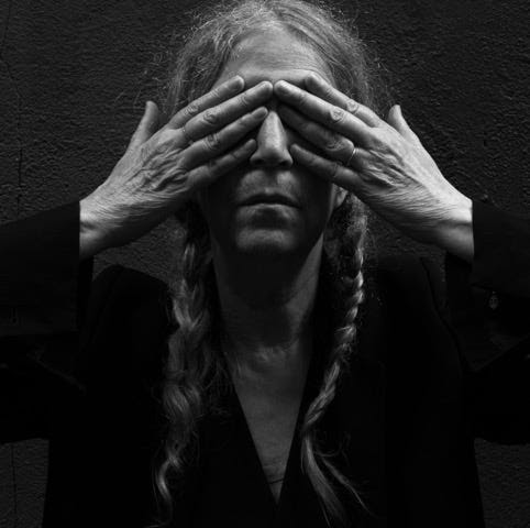 Portrait von Patti Smith in schwarz weiß, Sie hält sich beide Hände vor ihr Gesicht. Ihre Finger verdecken ihre Augen, Nase und Mund sind erkennbar. Ihre Haare sind zu zwei Zöpfen geflochten welche jeweils seitlich rechts und links über ihrem Schlüsselbein hängen.