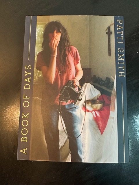 Buchcover der Taschenbuchausgabe von Patti Smith A Book Of Days. Das Bild ist von Patti Smith selbst aufgenommen und zeigt sie in rotem Oberteil und blauer Jeans. Ihre Finger der rechten Hand hält sie vor ihren Mund. In ihrer linken Hand hält sie ihre Polaroid.