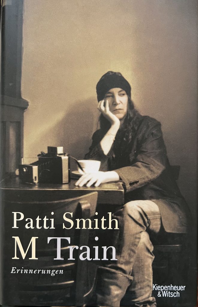 Patti Smith Buchcover M Train (deutsche Ausgabe, 2015). Das Foto des Covers zeigt Patti Smith sitzend an einer Art Sekretär, die Beine überschlagen, ihr Kopf mit ihrer rechten Hand stützend mit dem Ellenbogen auf dem Tisch, auf dem auch ihre Polaroid Land liegt.