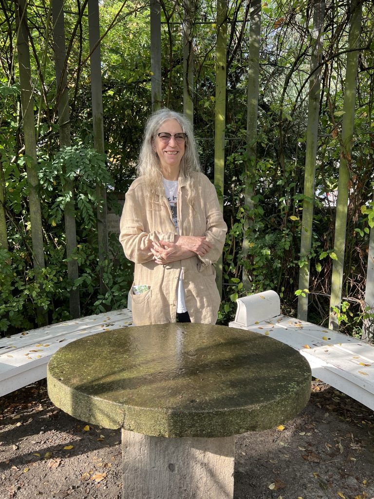 Bild von Patti Smith Besuch in Schillers magischem Garten in Jena, Deutschland im Oktober 2023. Das Bild zeigt Patti Smith hinter Schillers Tisch stehend, dabei schaut sie frontal in die Kamera und lächelt.