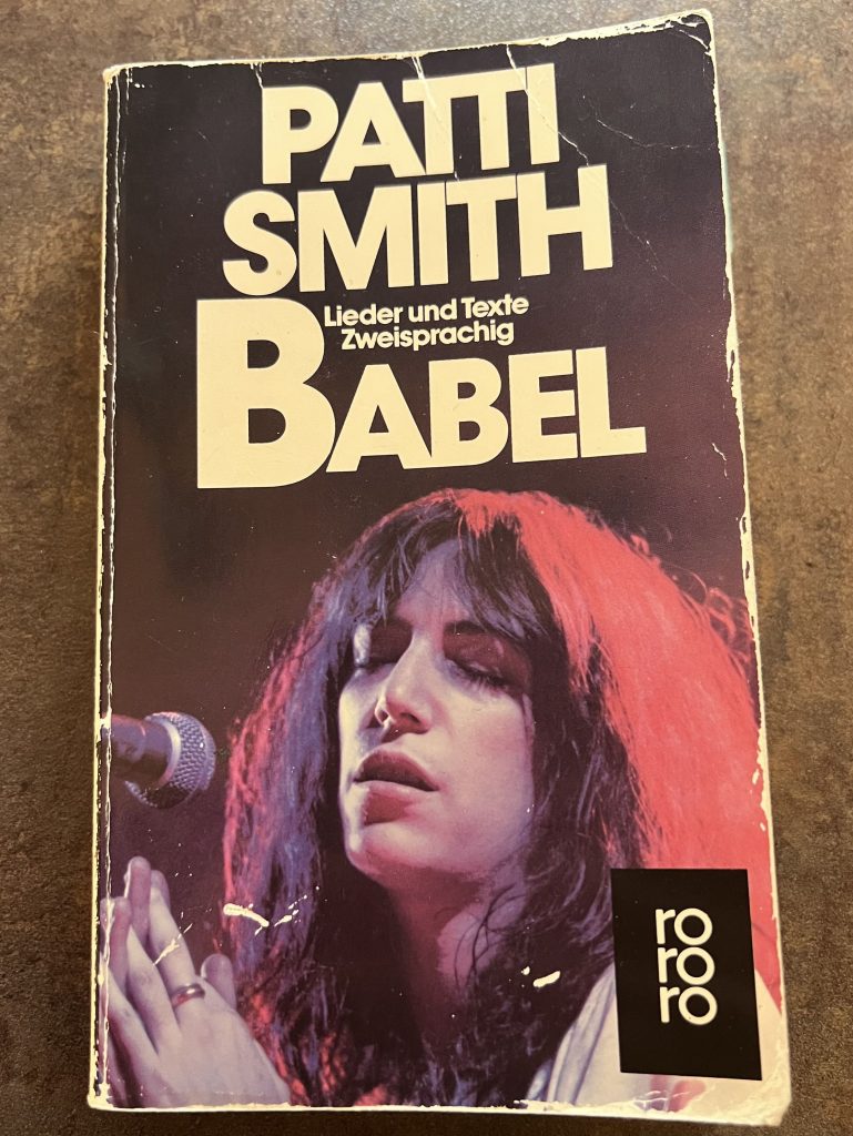 Buchcover der zweisprachigen Ausgabe von Patti Smith BABEL, welches 1985 veröffentlicht wurde. Es enthält ihre Gedichte, Prosa, Lyrik, Bilder und Zeichnungen in englisch und deutsch. Das Foto (Hans H. Kirmer) zeigt Patti Smith während eines Auftritts vor dem Mikrofon stehend in Naheaufnahme.