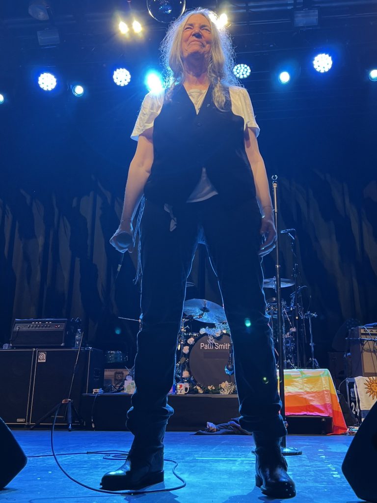 Patti Smith feiert ihren Geburtstag im Brooklyn Steel am 30. Dezember 2022. Das Bild zeigt Patti während ihrer Show auf der Bühne stehend. Kurz nach einem Song steht sie am Bühnenrand. Ihre Arme sind gerade nach unten, sie schaut leicht schmunzelnd ins Publikum.