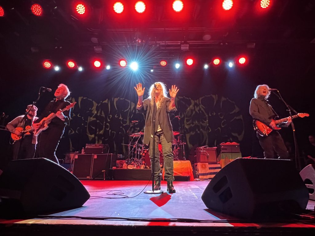Patti Smith feiert ihren Geburtstag im Brooklyn Steel am 30. Dezember 2022. Das Bild zeigt Patti in der Mitte auf der Bühne stehend, ihre Arme leicht nach oben während einer Performance. Zu ihrer Linken steht Lenny an der Gitarre, zu ihrer Rechten ihr Sohn Jackson an der Gitarre spielend und Tony Shanahan am Bass und singend.