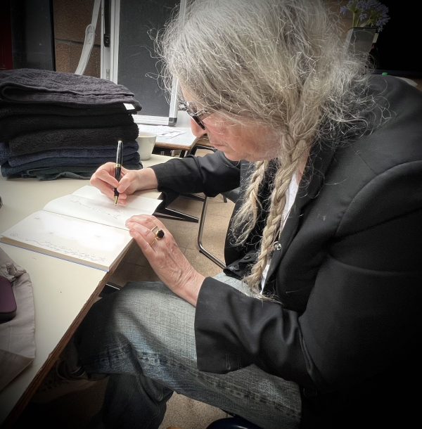 Patti Smith sitzt auf einem Stuhl am Tisch. Vor ihr ein aufgeschlagenes Buch. Links ist ein weiteres Design, vom Ersteller des Bildes unkenntlich gemacht, auf der rechten Seite zeichnet sie den "fugal fox compass". Sie trägt ein schwarzes Sakko, darunter ein weißes T-Shirt und eine blaue Jeans.