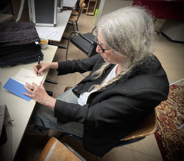 Patti Smith sitzt auf einem Stuhl am Tisch. Vor ihr ein aufgeschlagenes Buch. Links ist die Lasche des Einbandes, auf der rechten Seite zeichnet sie das erste Design des fugal fox. Sie trägt ein schwarzes Sakko, darunter ein weißes T-Shirt und eine blaue Jeans.