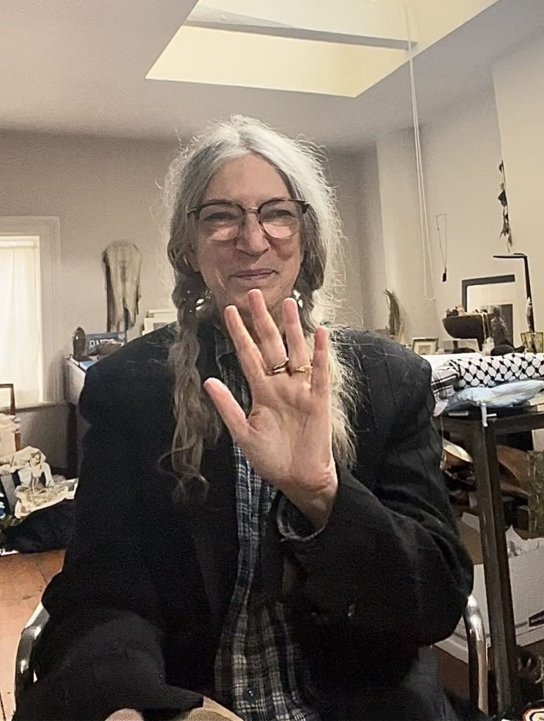 Patti Smith sitzt auf ihrem Stuhl und winkt mit ihrer linken Hand lächelnd in die Kamera. Ihre Haare sind zu zwei Zöpfen geflochten. Sie trägt ein schwarzes Sakko und darunter ein blau kariertes Hemd.