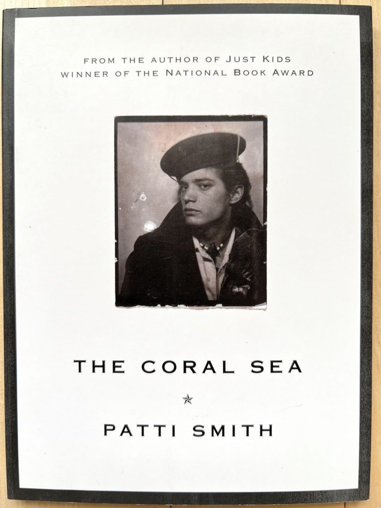 Das Buch "The Coral Sea" von Patti Smith liegend, mit der Vorderseite nach oben. Mittig ein Brustbild von Robert Mapplethorpe. Er trägt eine Mütze, eine Kette, eine Jacke mit hochgestelltem Kragen, darunter ein weißes Hemd. Darunter steht der Titel des Buches "The Coral Sea", darunter die Autorin Patti Smith