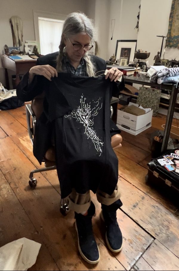 Patti Smith sitzt auf ihrem Stuhl und präsentiert die neuen fugal fox T-Shirts. Es sind zwei verschiedene Designs, jeweils in schwarz und weiß. Die Designs wurden von Patti selbst gezeichnet. Auf diesem Bild hält sie das schwarze T-Shirt mit dem ersten Design in die Kamera..