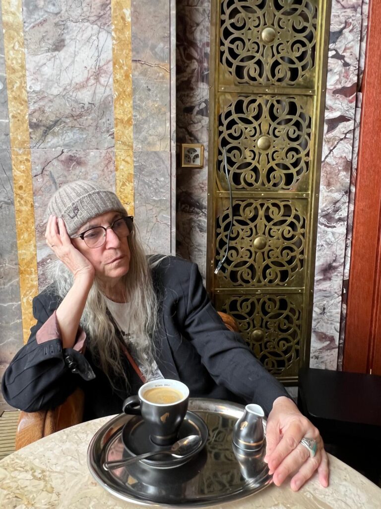 Patti Smith im Café Odeon in Zürich im Juli 2025 während ihrer Europa Tour. Patti sitzt an einem kleinen runden Tisch an einem Holzstuhl mit Armlehnen. Mit ihrem rechten Arm stützt sie ihren Kopf, ihre linke Hand hat sie auf dem Tisch abgelegt. Vor ihr steht ihr Café in einer schwarzen Julius Meinel Tasse auf einem silbernen kleinen Tablett zusammen mit einem kleinen Krug heißem Wasser.