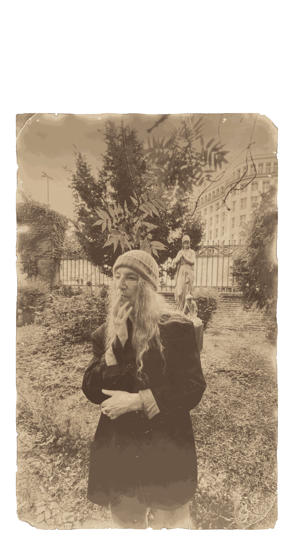 Patti Smith mit Strickmütze und dunklem Mantel in einem Garten in Berlin, die Hand am Kinn, hinter ihr Bäume und eine Steinfigur, 2025