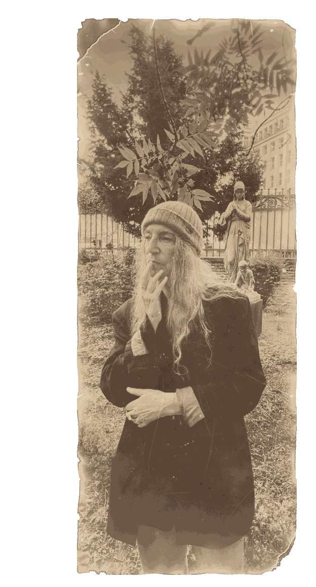 Patti Smith mit Strickmütze und dunklem Mantel in einem Garten in Berlin, die Hand am Kinn, hinter ihr Bäume und eine Steinfigur, 2025
