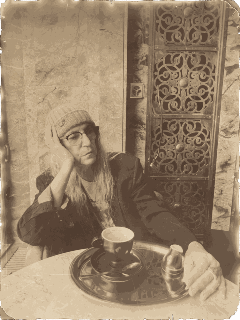Patti Smith mit Brille und Strickmütze im Café Odeon in Zürich, den Kopf in die Hand gestützt, vor ihr eine Tasse Kaffee auf einem Tablett, Juli 2025