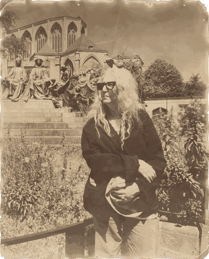 Patti Smith mit Sonnenbrille und einem Hut in der Hand vor einem Denkmal und einer gotischen Kirche in Gent, 2026
